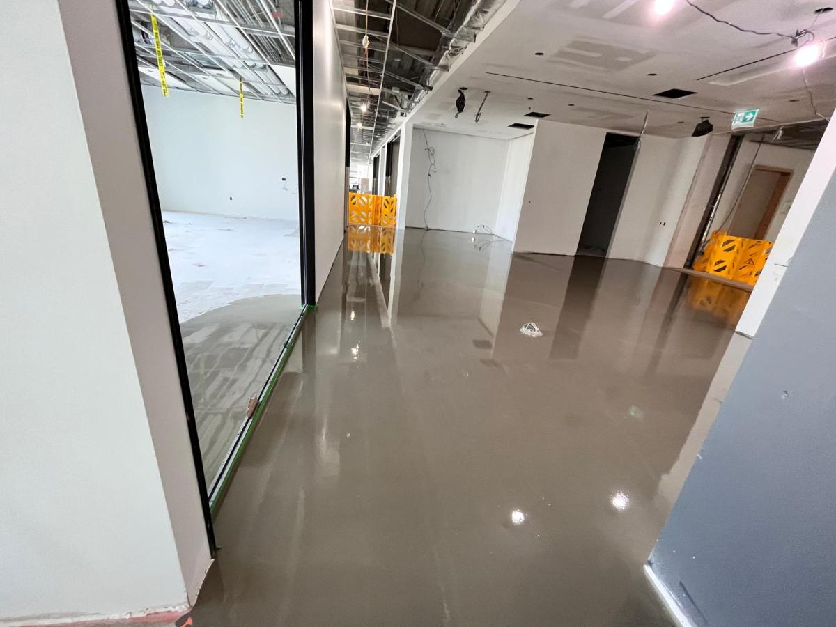 Self-levelling floor pour along a commercial corridor with black-framed glass partitions